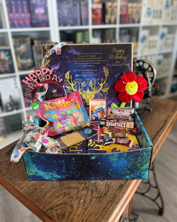 Das Produktbild zeigt vorn eine geöffnete Drachenpost in blauem Schuppen-Design. Man sieht den in den Boxdeckel geklebten Brief zum Thema #birthday und in der Box zum Thema passende Goodies - Süßigkeiten und andere Kleinigkeiten. Im Hintergrund sieht man die Bücherregale des Verlagsladens.